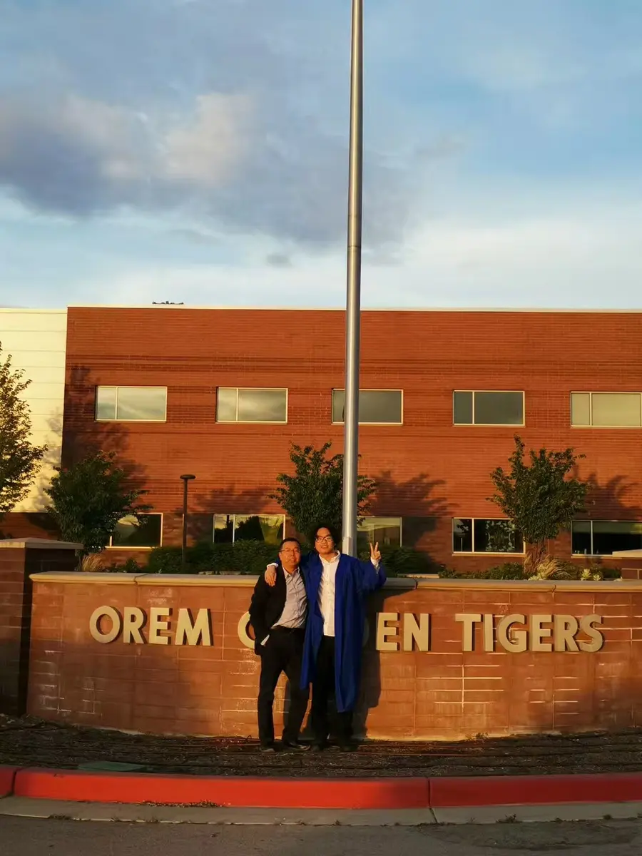 中国学生穿毕业服在 Orem High School 校名墙前与家人或教师合影